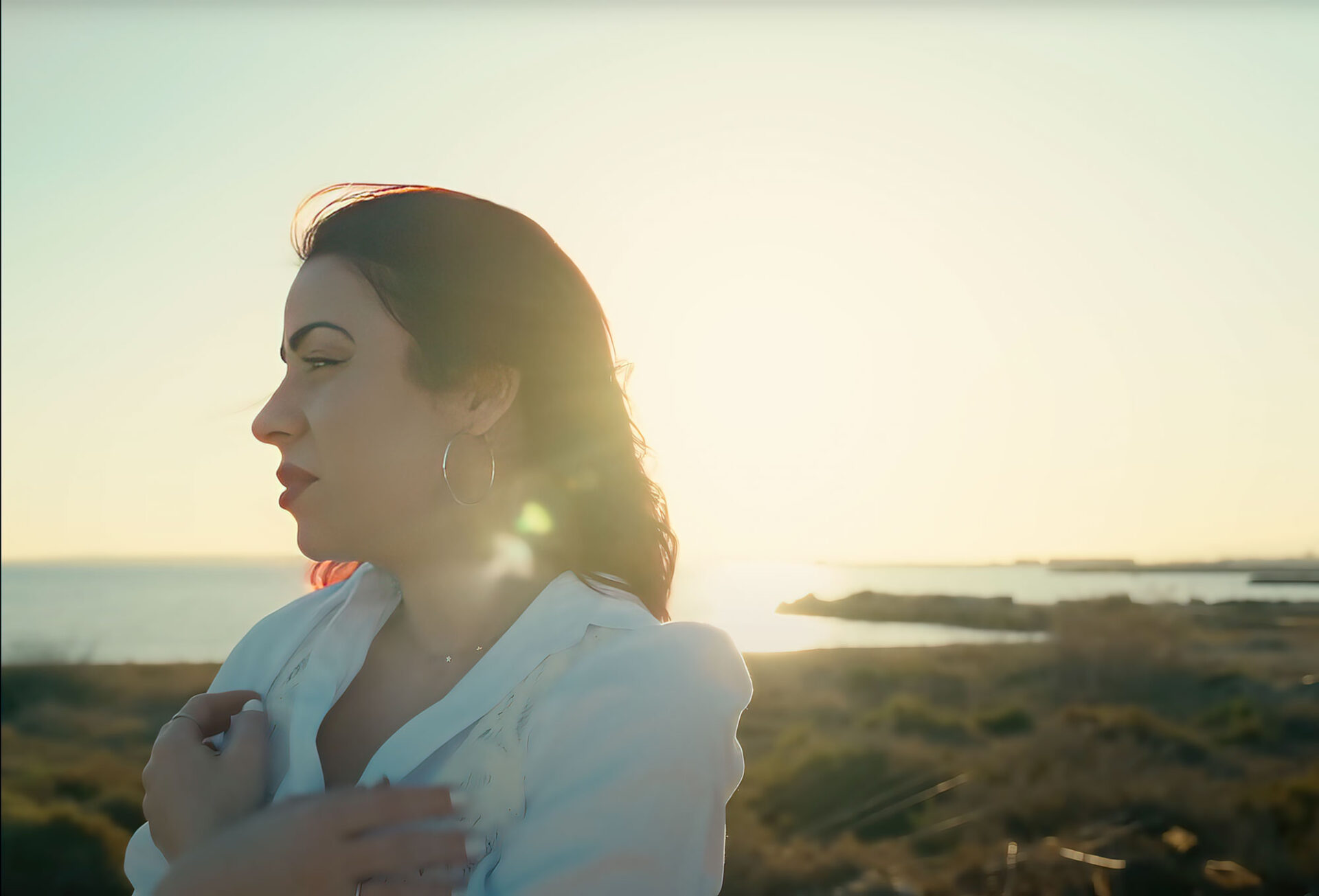 fotograma del videoclip de Fugaz, la canción debut de Verónica Terrés. Se ve en primer plano a Verónica, vestida con una camisa blanca, abrazándose a sí misma y mirando hacia la izquierda, y al fondo desenfocado se ve el atardecer en la playa junto al mar.